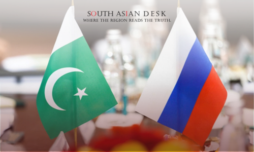 Two small flags, one of Pakistan with a green background, white stripe, crescent moon, and star, and the other of Russia with white, blue, and red vertical stripes, placed on a table with blurred bottles and decorations in the background.