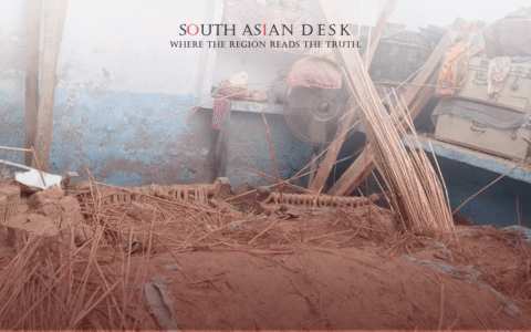 A scene of destruction with scattered wooden sticks, debris, and mud in front of a blue and white wall. Items such as a fan, suitcases, and colorful fabrics are visible among the wreckage.