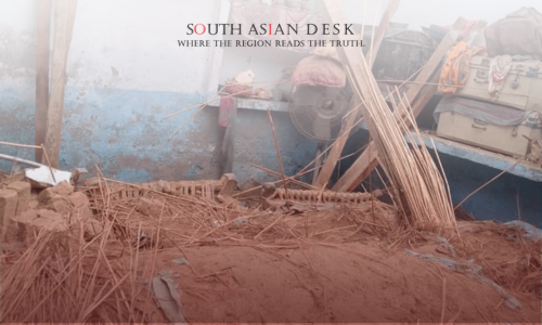 A scene of destruction with scattered wooden sticks, debris, and mud in front of a blue and white wall. Items such as a fan, suitcases, and colorful fabrics are visible among the wreckage.