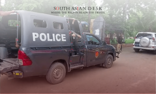 A police truck with 'POLICE' written on the side, parked on a dirt road with officers exiting, surrounded by trees and a gated area, with another vehicle in the background.