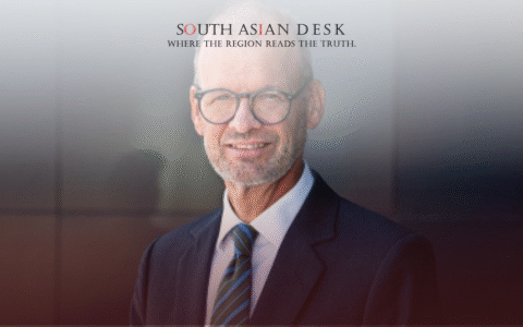 A man in a dark suit and tie with glasses, smiling against a dark, reflective background.