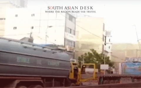 A large water tanker truck driving on a road with buildings and other vehicles in the background.