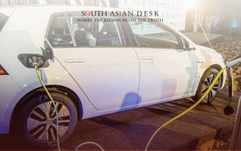A white electric car connected to a charging station with a yellow cable, displayed at an event with a backdrop featuring logos.