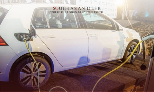 A white electric car connected to a charging station with a yellow cable, displayed at an event with a backdrop featuring logos.