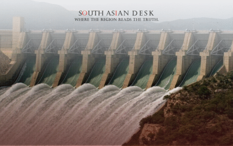 A large dam with multiple spillways releasing water, surrounded by hilly terrain and greenery.