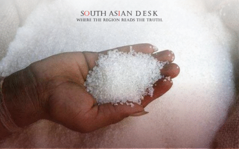 A hand holding a pile of white sugar crystals, with a large sack of sugar in the background.