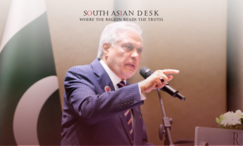 A man in a suit speaking at a podium with a microphone, gesturing with his hand, with the Pakistani flag and a floral arrangement in the background.