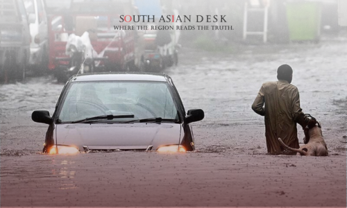 A car with its headlights on is partially submerged in floodwater on a street, while a person in a brown outfit stands in the water beside a dog, with other vehicles and a green bin visible in the background.