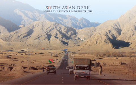 A desert highway flanked by rugged mountains, with several vehicles and a person walking along the road.
