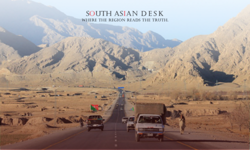 A desert highway flanked by rugged mountains, with several vehicles and a person walking along the road.