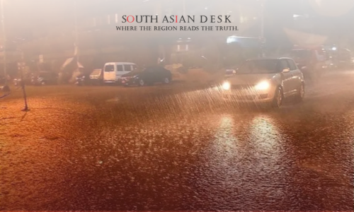 A car driving through a flooded street at night, with water splashing around its tires, under the glow of streetlights and surrounded by other vehicles.