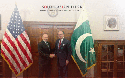 Two men in suits shaking hands in front of the American and Pakistani flags, with a wooden door and official decor in the background.