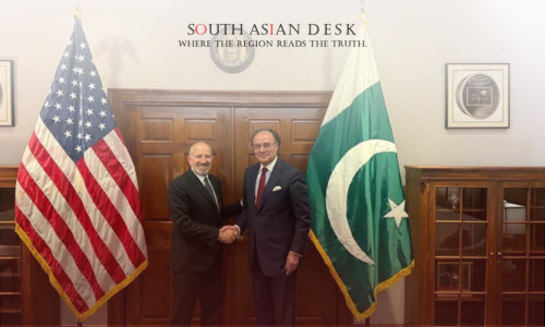 Two men in suits shaking hands in front of the American and Pakistani flags, with a wooden door and official decor in the background.