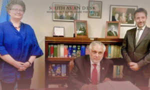A woman in a blue outfit and two men in suits standing in an office with a bookshelf, framed photos, and a flag in the background, with one man signing a document.