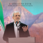 A man in a suit speaking at a podium with microphones, standing in front of an Iranian flag and a mountainous backdrop.