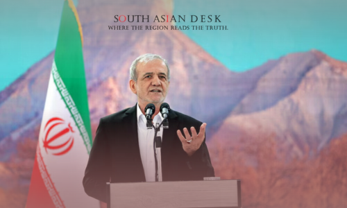 A man in a suit speaking at a podium with microphones, standing in front of an Iranian flag and a mountainous backdrop.