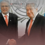 Two men in suits shaking hands in front of UN flags and a backdrop with the UN logo.