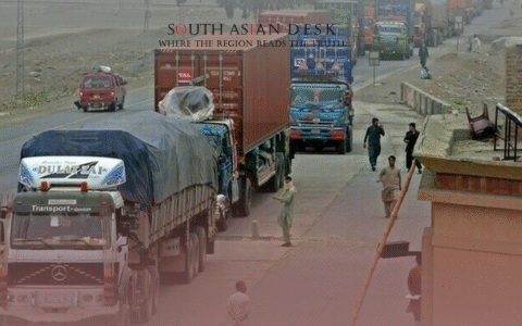 A busy road scene with multiple trucks and containers lined up, people walking alongside, and a rural or semi-urban setting with a checkpoint or border-like atmosphere.