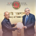 Two individuals in a formal setting, exchanging a decorative plate, with a backdrop featuring the emblem and text of the Ministry of Home Affairs in Bengali.