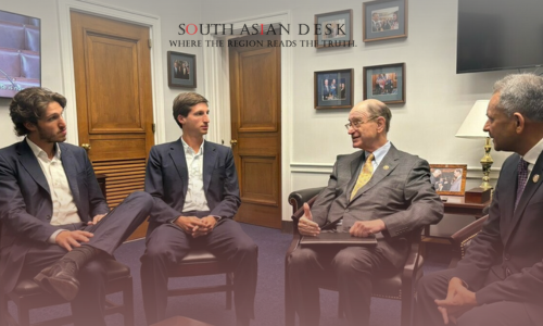 Four men in suits sit in a formal office setting with wooden doors, framed photos, and a television on the wall. The man on the right, an official with a pin, and the man in the center with a yellow tie, a U.S. politician, engage in discussion, while two younger men listen attentively.