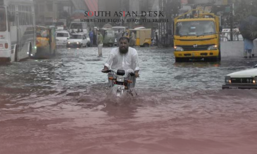 A man in a white outfit rides a motorcycle through a flooded urban street, with water splashing around the wheels. In the background, a yellow truck, a white bus, a car, and other vehicles are partially submerged, while people stand on the sidewalks under a gray sky.