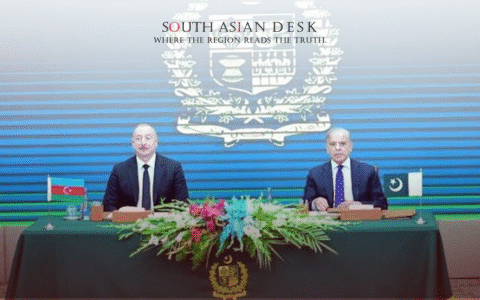 Two men in suits sitting at a table covered with a green cloth, with a large floral arrangement in the center, during a formal meeting. The backdrop features the emblem of Pakistan, and small flags of Azerbaijan and Pakistan are placed on the table.