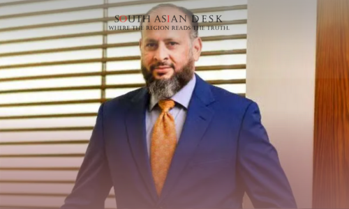 A man in a blue suit with an orange tie standing indoors, with wooden blinds and a wooden wall in the background.