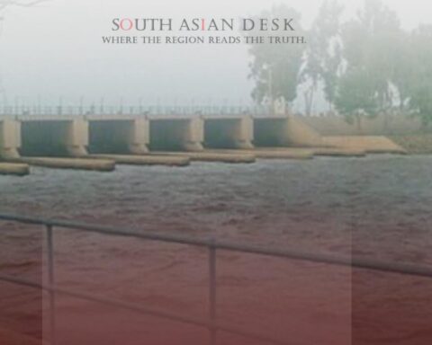 A dam or barrage structure with water flowing through multiple gates, surrounded by a railing and trees in the background.