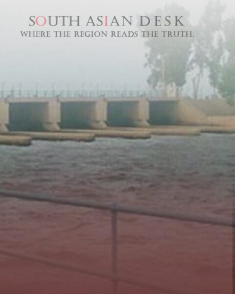A dam or barrage structure with water flowing through multiple gates, surrounded by a railing and trees in the background.