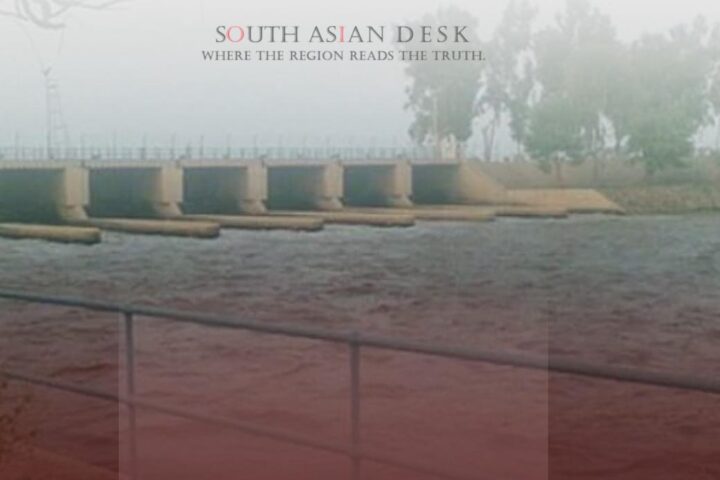 A dam or barrage structure with water flowing through multiple gates, surrounded by a railing and trees in the background.