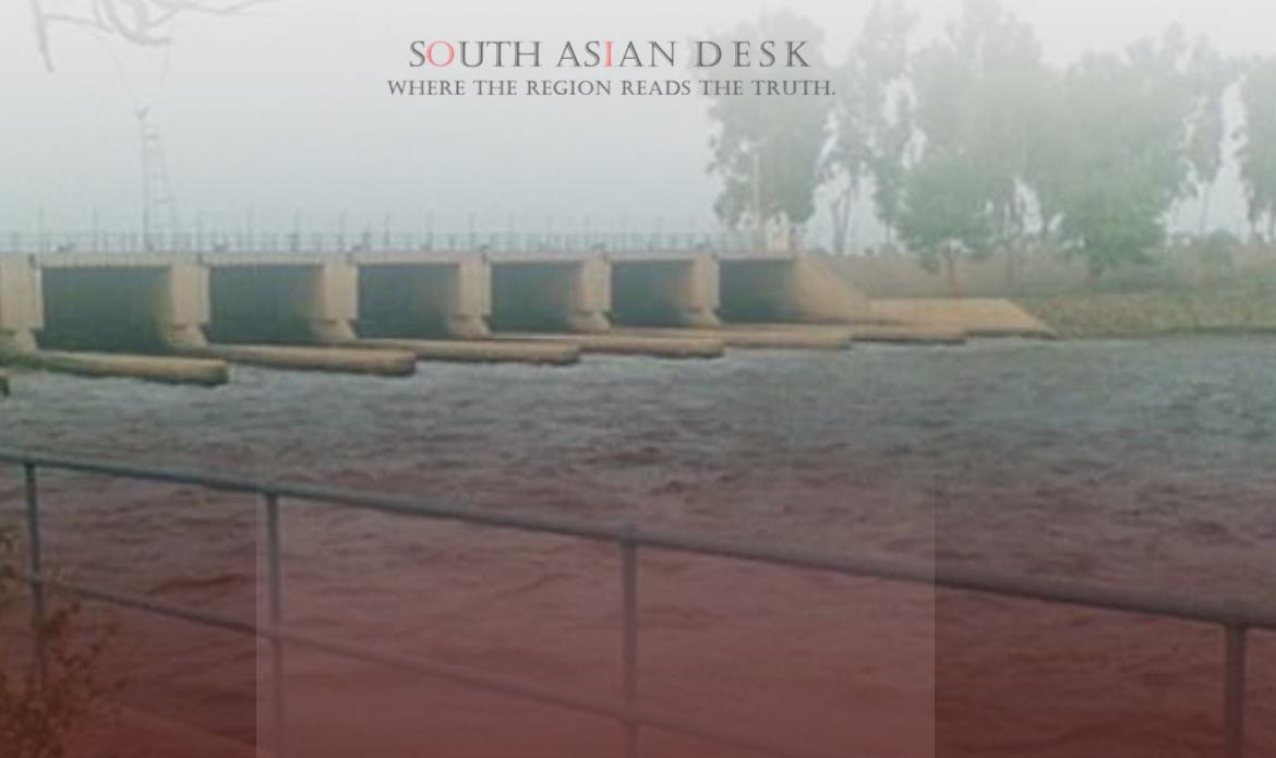 A dam or barrage structure with water flowing through multiple gates, surrounded by a railing and trees in the background.