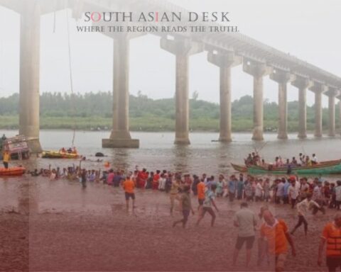 A partially collapsed bridge over a river with rescue boats and people in orange life jackets assisting near the water and muddy shore.