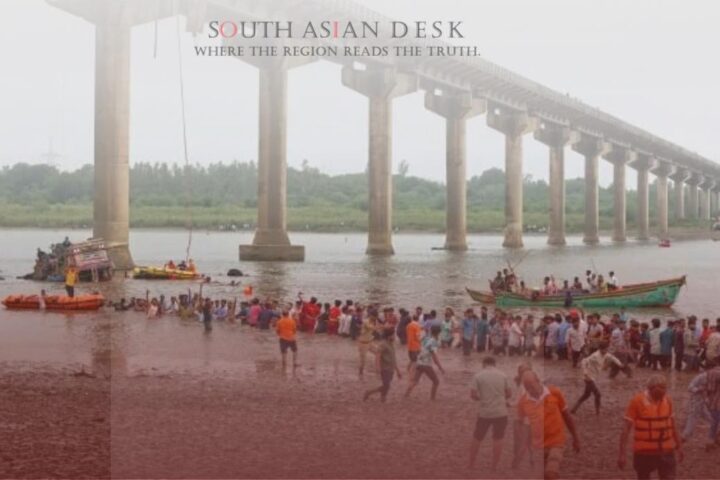 A partially collapsed bridge over a river with rescue boats and people in orange life jackets assisting near the water and muddy shore.
