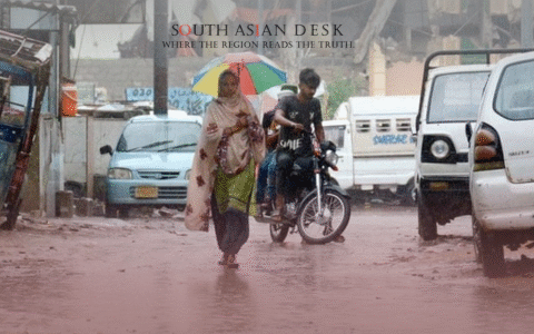 A woman holding a colorful umbrella walks on a wet street during rainfall, with a motorcyclist and parked cars in the background, suggesting an urban setting with some structural damage, depicting longer monsoon in Pakistan.
